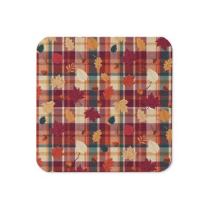 May include: A square coaster featuring a plaid pattern in shades of burgundy, orange, and cream. The surface is decorated with a repeating print of various autumn leaves in red, yellow, and brown, along with small brown acorns scattered throughout.