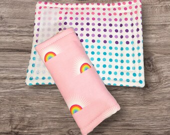May include: A pink fabric glasses case with a rainbow and sunburst pattern, next to a white square coaster with a polka dot pattern in shades of pink, purple, and blue. The items are on a wooden surface.