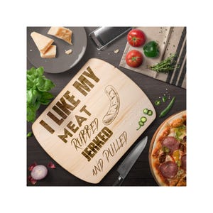 May include: A wooden cutting board with the text "I like my meat rubbed, jerked and pulled" engraved on it. The board is surrounded by various kitchen items, including a pizza, cheese, and herbs.