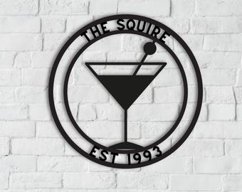 May include: Black metal sign with the words "THE SQUIRE EST 1993" in a circular design. A martini glass is in the center of the circle. The sign is mounted on a white brick wall.