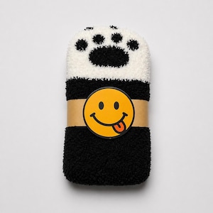 May include: A pair of fuzzy black and white socks featuring a paw print design on the top section. The socks are folded and secured with a tan paper band that displays a yellow circular smiley face icon with a tongue sticking out.