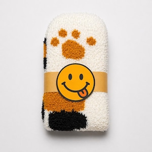 May include: A pair of folded fuzzy socks featuring a white base with black and orange patches. The socks are wrapped in a tan paper band displaying a yellow circular smiley face icon with a tongue sticking out, centered on the front.
