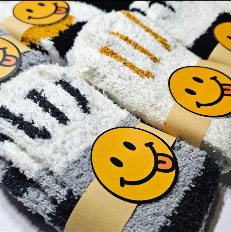 May include: A pair of fuzzy, plush socks featuring a white and gray color scheme with black accents. The socks are wrapped in a tan paper band decorated with a yellow circular smiley face icon that has a tongue sticking out to the side.