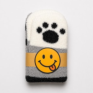 May include: A pair of folded fuzzy socks featuring a white upper section with a black paw print design, a grey middle section, and a black toe section. A tan paper band wraps around the center, displaying a yellow smiley face with a tongue out.