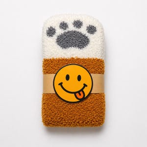 May include: A pair of folded fuzzy socks featuring a white top section with a grey paw print design and a solid mustard yellow bottom section. A tan paper band wraps around the center, displaying a yellow smiley face icon with a tongue out.