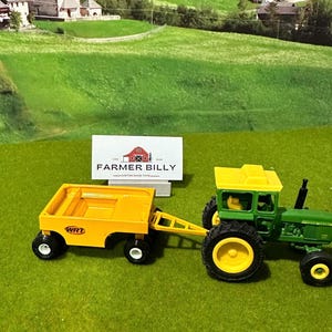May include: A green toy tractor with a yellow cab and a yellow wagon attached. The wagon has the text "WRT" on the side. The tractor and wagon are on a green surface.