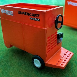 May include: A bright orange miniature Valmetal Supercart VA 542 agricultural feed cart model. The toy features a black steering wheel, a perforated front platform, and a single black wheel with a white center hub, displaying clear black text branding.