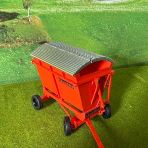 May include: A bright orange toy farm trailer with a silver corrugated roof and black wheels. The trailer has the name "BICKMPTON" printed on the sides. The background features a green field and a church.