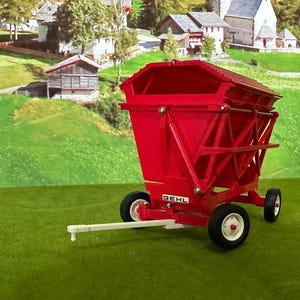 May include: A bright red toy farm wagon with black tires and white rims. The wagon has a rectangular body and is set against a backdrop of a green field and rural buildings.