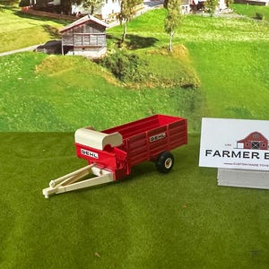 May include: A red and cream toy farm trailer with a single black tire. The trailer has the word "GEHL" printed on the side. A white sign with the words "FARMER BILLY" is to the right.