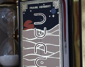 Könnte beinhalten: Ein Hardcover-Buch mit schwarzem Einband und goldenen Lettern. Der Titel des Buches lautet "Dune" von Frank Herbert. Der Einband zeigt ein stilisiertes Bild einer Sanddüne mit einer roten Sonne, die im Hintergrund untergeht.