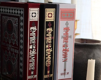 May include: A stack of three hardcover books with gold lettering and decorative designs. The books are titled "Empire of the Vampire", "Empire of the Damned", and "Empire of the Dawn" by Jay Kristoff. The books are in a dark setting with candles.