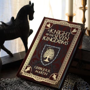 Puede incluir: Un libro de tapa dura burdeos titulado "A Knight of the Seven Kingdoms" con letras doradas y bordes decorativos. El libro presenta un emblema de escudo. También se ven una estatua de caballo negro, una espada y cota de malla.