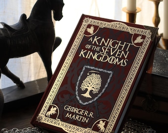 Puede incluir: Un libro de tapa dura burdeos titulado "A Knight of the Seven Kingdoms" con letras doradas y bordes decorativos. El libro presenta un emblema de escudo. También se ven una estatua de caballo negro, una espada y cota de malla.
