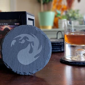 May include: A round, dark gray slate coaster with a white, stylized flame design. A glass of amber liquid sits nearby on a matching coaster. A black card holder with a Magic: The Gathering card is in the background.