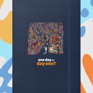 May include: A navy blue journal with an abstract design and the text "one day or day one?". The design features colorful lines and shapes. The journal has an elastic band closure.