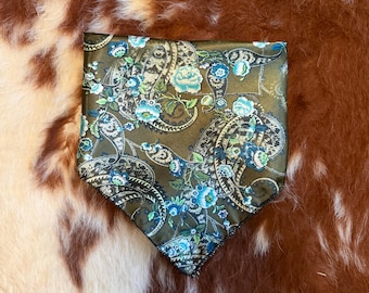 May include: A square, olive green pocket square with a detailed paisley and floral pattern in shades of blue, teal, and white. The edges are finished with a dark blue border. The fabric has a slight sheen.