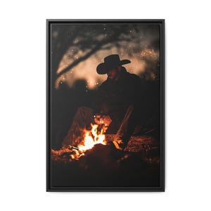 May include: A framed canvas print depicts a person in a cowboy hat sitting by a campfire at dusk. The fire's orange and yellow flames illuminate the scene, with sparks and a blurred background of trees and sky.