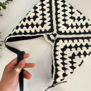 May include: A triangular head covering crafted from black and white yarn in a classic granny square crochet pattern. The accessory features a solid black fabric lining and includes long, thin black straps attached to the bottom corners for tying.