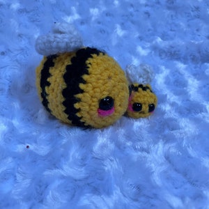 May include: Two crocheted bee plushies, one larger than the other, with black and yellow stripes and white wings. The larger bee has pink cheeks and black eyes.