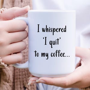 May include: White ceramic coffee mug with the text "I whispered 'I quit' to my coffee..." in black script. The mug is held by a person wearing a beige jumper. The mug has a large handle.