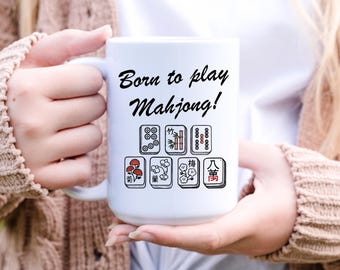 May include: White ceramic mug with the text "Born to play Mahjong!" in black script. The mug features various Mahjong tile designs in red, black, and green. The mug is held by a person.