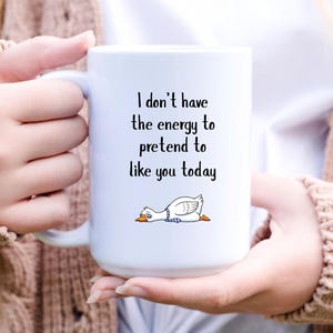 以下が含まれることがあります： 「I don't have the energy to pretend to like you today」の文字とアヒルのイラストが描かれた白いセラミックマグカップ。アヒルは白で、オレンジ色のくちばしと足があり、横たわっています。マグカップは人が持っています。