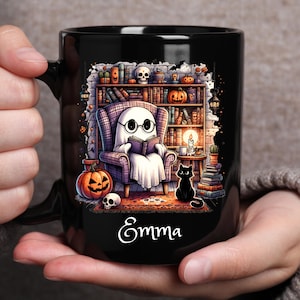 May include: Black ceramic mug featuring a Halloween-themed illustration. A ghost wearing glasses reads in a cozy armchair in front of a bookshelf. A jack-o'-lantern, skull, and black cat are also depicted. The name "Emma" is written on the mug.
