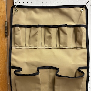 May include: Tan tool organizer with multiple pockets and black trim. The organizer has several small pockets at the top and three larger pockets at the bottom. It is designed to hang on a wall or pegboard.