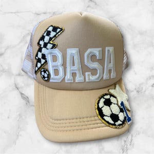 May include: A tan baseball cap with a white mesh back. The cap has the word "BASA" in white block letters, a black and white checkered lightning bolt, and soccer ball and star patches. The brim is tan.