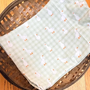 May include: A light green and white gingham fabric featuring a repeating pattern of small white geese with orange beaks and feet. The fabric is folded and draped over a round, dark brown woven basket with a lattice design.