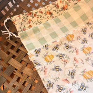 May include: Three fabric bags with drawstring closures rest in a woven basket. The bags feature different patterns: floral, gingham, and a bee-themed design with honeycombs and flowers. The color palette includes cream, green, pink, and yellow.