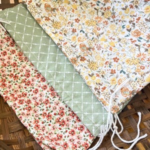 May include: Three drawstring bags with floral patterns. One bag features a pattern of red flowers, another has a green diamond pattern, and the third has a yellow and orange floral design. The bags are made of fabric and have white drawstrings.