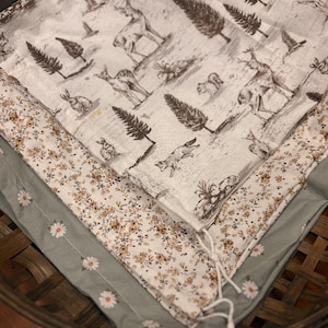 May include: A stack of three fabric layers with different patterns. The top layer is white with a sepia-toned forest scene featuring deer, rabbits, and trees. The middle layer has a floral pattern in cream and brown. The bottom layer is sage green with white daisy accents.