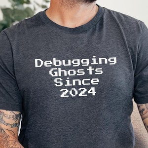 Könnte beinhalten: Ein dunkelgraues T-Shirt mit dem Text "Debugging Ghosts Since 2024" in weißer Pixel-Schrift.