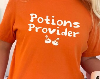 May include: Orange t-shirt with the text "Potions Provider" in white, featuring two potion bottle graphics. The shirt is part of a Halloween theme, with a decorative pumpkin and ghost-themed decor in the background.