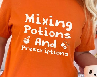 May include: Orange t-shirt with the text "Mixing Potions And Prescriptions" in white. The shirt is worn by a person holding a white mug with a ghost design and a candy cane.