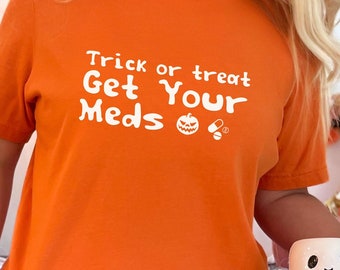 May include: An orange short-sleeved t-shirt featuring white text that reads Trick or treat Get Your Meds, accompanied by a small graphic of a jack-o'-lantern and two pills. The shirt has a crew neckline and a standard fit.