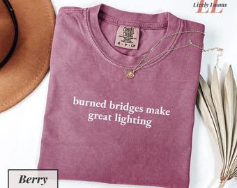 Puede incluir: Camiseta de color bayas con el texto blanco "burned bridges make great lighting". La camiseta tiene cuello redondo y mangas cortas. Un collar dorado con colgante está sobre la camiseta.