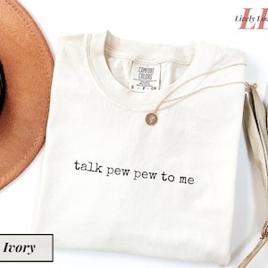 Peut inclure: T-shirt ivoire avec l'inscription noire "talk pew pew to me". Un collier doré avec un pendentif est posé sur le t-shirt. Le t-shirt a une étiquette Comfort Colors.