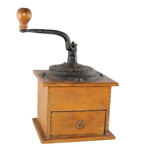 May include: A manual coffee grinder with a square light brown wooden box base featuring a small front drawer with a circular knob. The top assembly consists of a dark cast iron mechanism with an ornate embossed pattern and a curved handle.