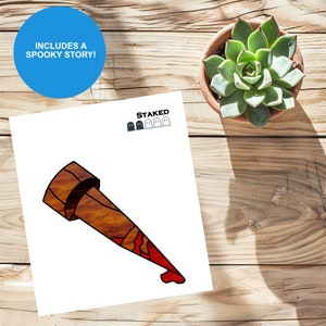 May include: A wooden stake with a red drip of blood at the tip. The stake is a Halloween decoration and includes a spooky story. The text "STAKED" is printed above the stake with five ghost icons.