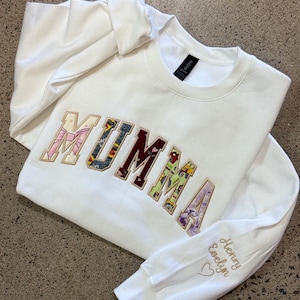 May include: Cream-coloured sweatshirt featuring the embroidered word "MUMMA" in a patchwork design. The sleeve has the embroidered names "Henry Evelyn" and a heart symbol. The sweatshirt is made of a soft material and is a comfortable fit.