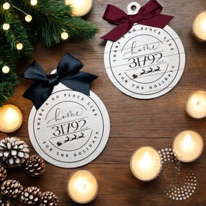 Puede incluir: Dos adornos navideños redondos de madera con el texto "There's no place like home 31792 for the holidays". Uno tiene un lazo negro, el otro un lazo burdeos. Están sobre una superficie de madera con velas y piñas.