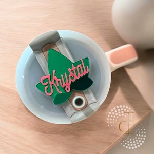 Puede incluir: Un adorno de árbol de Navidad de acrílico verde y rosa con el nombre "Krystal" escrito en él. El adorno está sentado en un plato de cerámica blanco con un asa rosa.