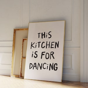 Puede incluir: Un lienzo blanco con un marco dorado apoyado contra una pared blanca. El lienzo tiene un texto escrito a mano en negro que dice "This kitchen is for dancing."
