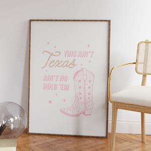 May include: A framed print with a pink cowboy boot and the text "This ain't Texas ain't no hold 'em" in a pink script font. The print is surrounded by a brown frame and is hanging on a white wall.