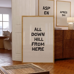 May include: A framed print with the text "ALL DOWN HILL FROM HERE" in black lettering on a white background. The print is hanging on a wall in a room with a wooden floor and a wooden dresser.
