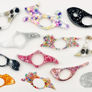 May include: A collection of 12 colourful and unique bookmark designs. Each bookmark features a different design, including a cat, a whale, a dog, and various abstract patterns. The bookmarks are made of clear resin and are filled with glitter, seashells, and other decorative elements.