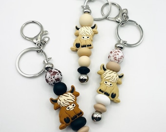May include: Three keychains with brown, white, and black beads and a small highland cow charm. The keychains are made of metal and have a silver finish.